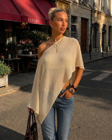 A woman stands on a city street wearing a lightweight, asymmetrical one-shoulder knit top in a warm cream or oatmeal color, paired with blue jeans. The background features a street scene with a cafe awning reading 'FLEURS'.