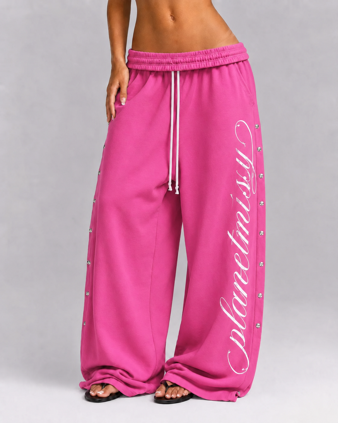 Bright pink wide-leg sweatpants with white script text reading 'planetisy' down one leg, white drawstrings, and small silver studs along the side seams, shown on a model against a light gray studio background.