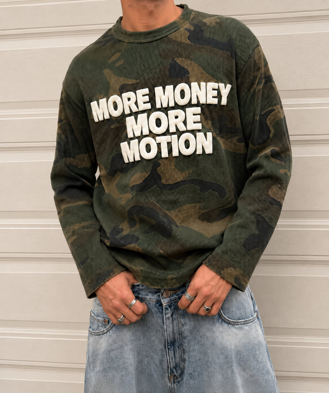 A person wearing a long-sleeve camouflage sweatshirt with the bold white text 'MORE MONEY MORE MOTION' across the chest, paired with light blue jeans, standing against a light beige paneled wall.