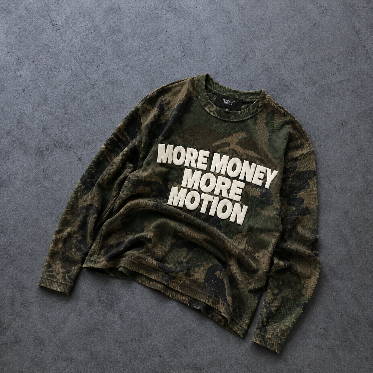 A long-sleeve crewneck sweatshirt in a green and brown camouflage pattern with the text 'MORE MONEY MORE MOTION' printed in bold white letters across the chest, laid flat on a textured gray concrete surface.