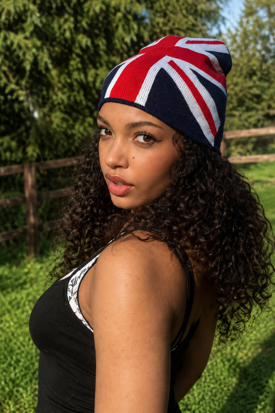 A woman outdoors wearing a beanie with a Union Jack flag pattern, standing in a grassy field with trees and a fence behind her.