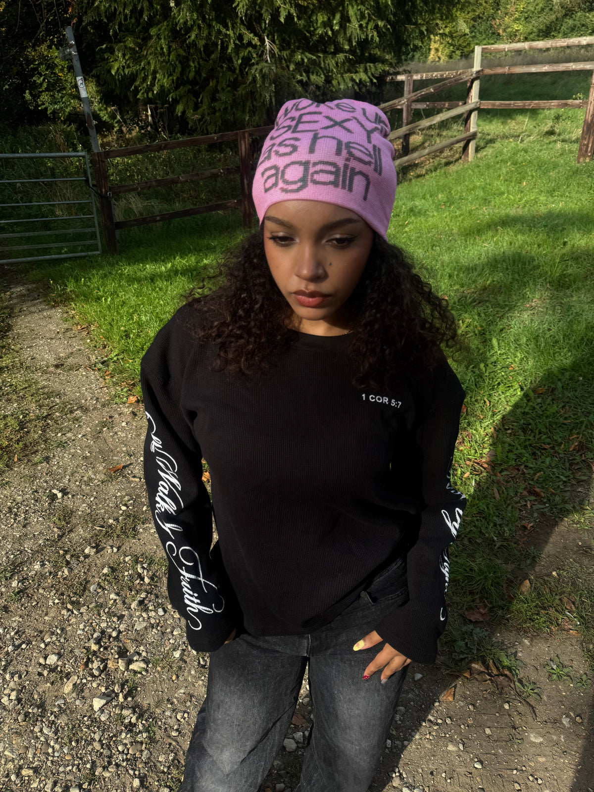 A person standing outdoors on a gravel path wearing a pink beanie with black text that reads 'SEX IS hell again' and a black long-sleeve shirt with white script graphics on the sleeves.