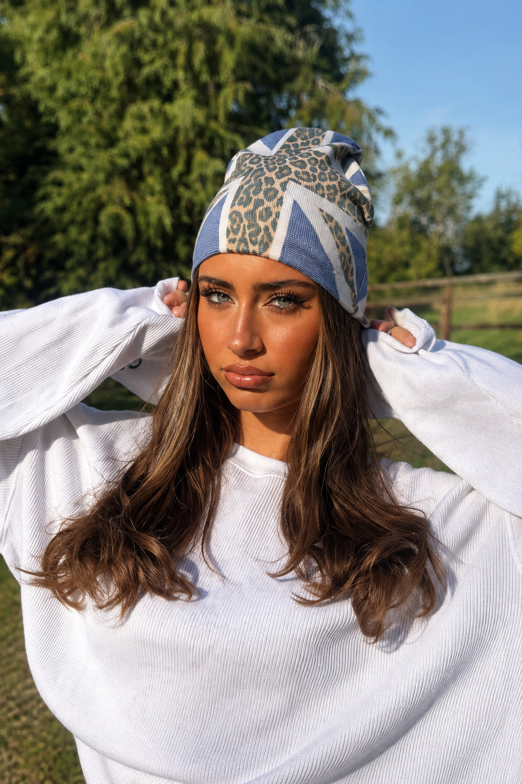 A woman wearing a white oversized crewneck sweatshirt, black athletic shorts, and a blue and white patterned beanie stands outdoors on a gravel path with trees and a fence in the background.
