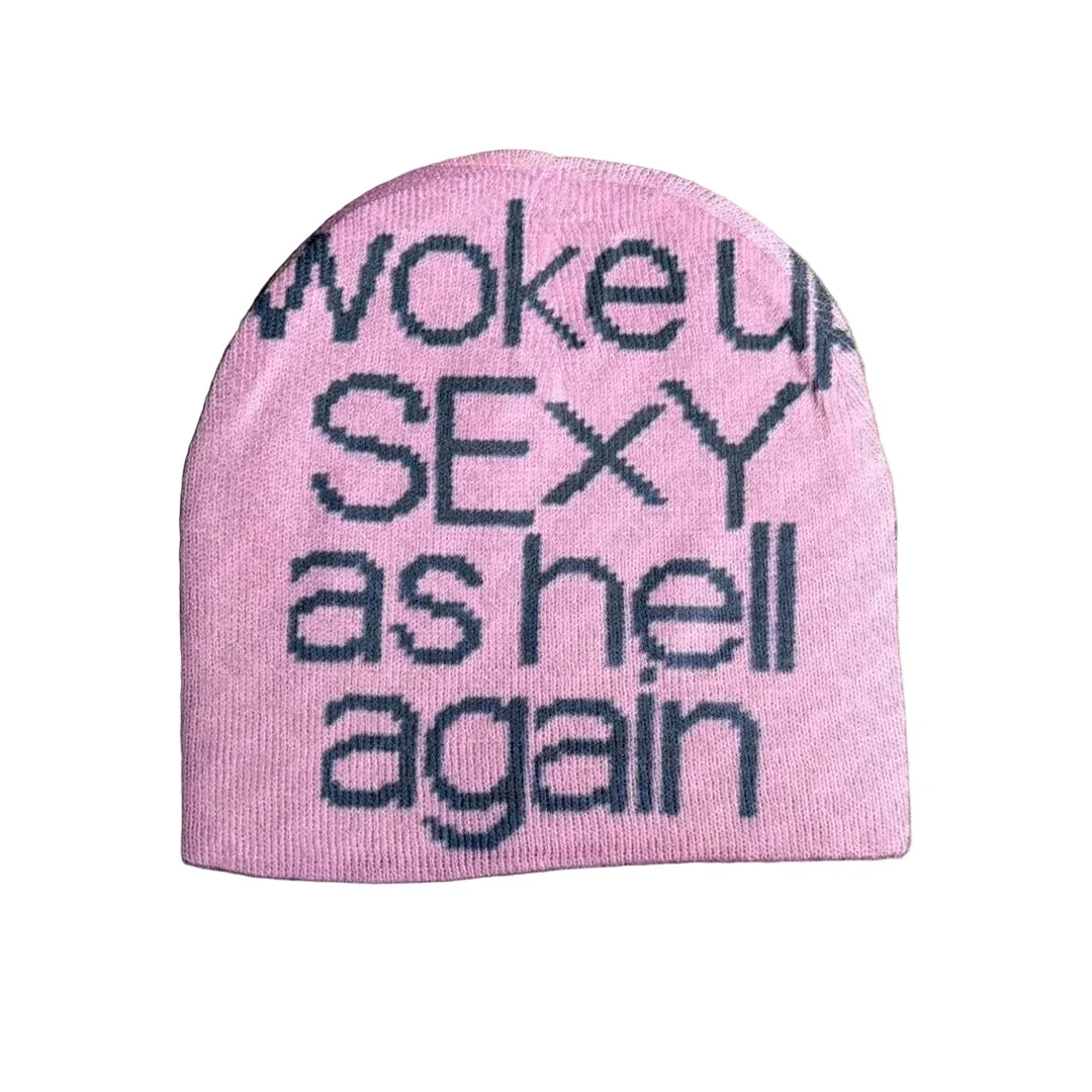 A pink knit beanie hat with the phrase 'woke up SEXY as hell again' knitted in dark navy lettering, laid flat on a textured gray concrete surface.