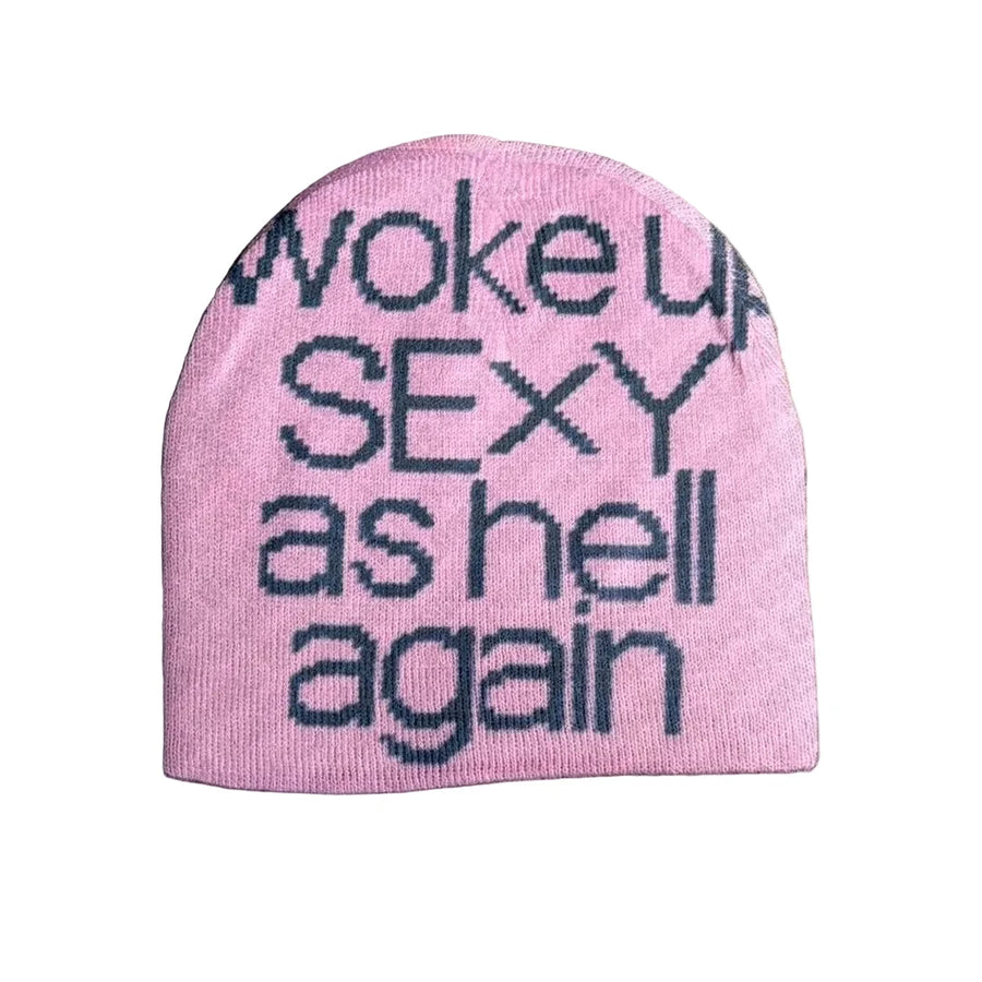 A pink knit beanie hat with the phrase 'woke up SEXY as hell again' knitted in dark navy lettering, laid flat on a textured gray concrete surface.