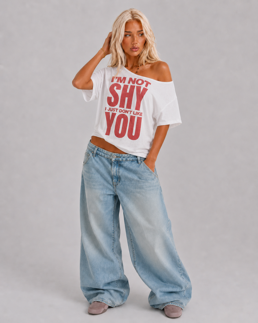 A blonde model wearing a white off-the-shoulder graphic t-shirt with red text that reads 'I'M NOT SHY I JUST DON'T LIKE YOU', paired with light-wash wide-leg jeans and gray slip-on shoes, against a neutral gray studio background.