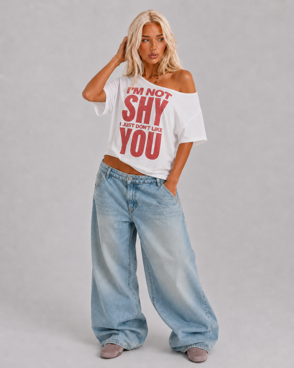 A blonde model wearing a white off-the-shoulder graphic t-shirt with red text that reads 'I'M NOT SHY I JUST DON'T LIKE YOU', paired with light-wash wide-leg jeans and gray slip-on shoes, against a neutral gray studio background.