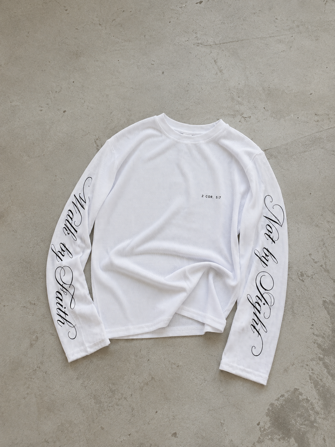 A white long-sleeve t-shirt with black cursive text printed down both sleeves, laid flat on a textured gray concrete surface.