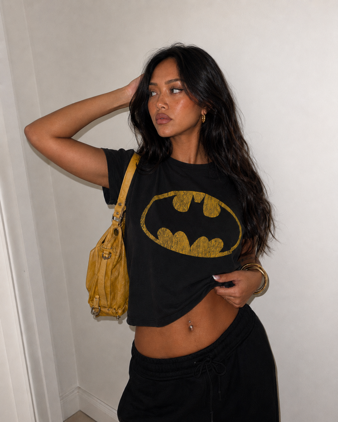 A woman posing in a black cropped t-shirt with a gold Batman logo, paired with black sweatpants and a yellow shoulder bag, set against a plain white wall.